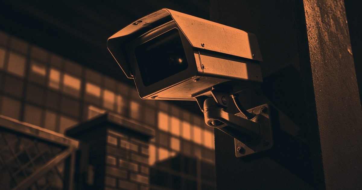 A CCTV camera on a post beside a brick gate pier and railings in a charcoal and orange duotone